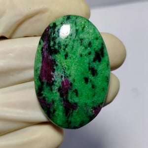 Puede incluir: Gema ovalada con una base verde vibrante, salpicada de inclusiones negras y rojo intenso. La superficie pulida refleja la luz, resaltando los patrones y texturas naturales de la piedra. Ideal para la creación de joyas.