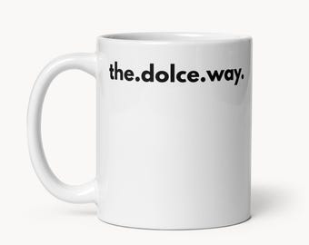 May include: White ceramic coffee mug with a curved handle. The mug features the phrase "the.dolce.way." printed in black, centered on the front. The mug is set against a plain, light background.