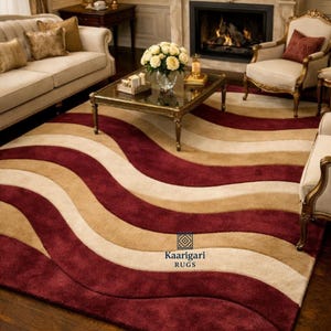 Puede incluir: Una alfombra grande con un patrón ondulado en colores burdeos, beige y crema. La alfombra está colocada en un salón con un sofá, sillas y una chimenea. La alfombra tiene el texto "Kaarigari RUGS".