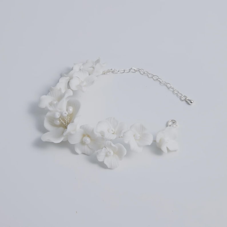 May include: A white floral bracelet with a silver chain and clasp. The bracelet features multiple white flower blossoms with pearl accents. The jewelry is designed for a delicate and feminine aesthetic.