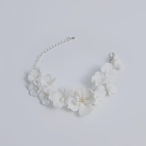 May include: A white floral bracelet with delicate, sculpted flowers and small pearl accents. The bracelet features a silver chain and clasp for adjustable sizing. The flowers are clustered together, creating a beautiful, elegant design.