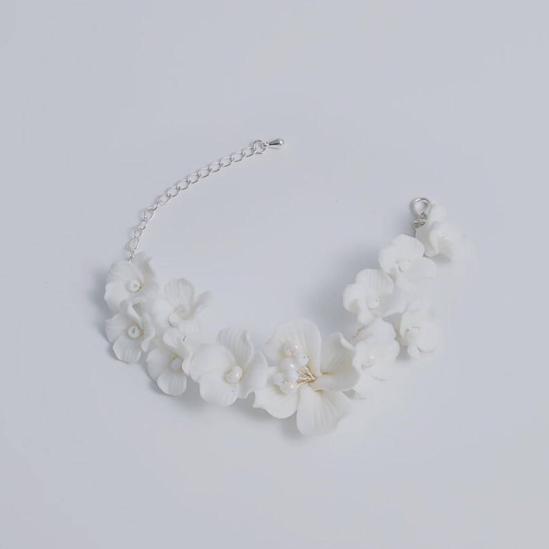 May include: A white floral bracelet with delicate, sculpted flowers and small pearl accents. The bracelet features a silver chain and clasp for adjustable sizing. The flowers are clustered together, creating a beautiful, elegant design.