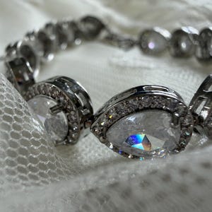 May include: A silver-toned link bracelet featuring a series of pear-shaped clear stones, each surrounded by a halo of smaller round clear stones. The metal settings are polished and reflective, showcasing a faceted design that catches the light.