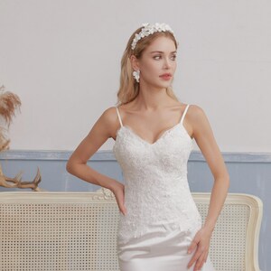 May include: A white wedding dress with a V-neckline and spaghetti straps. The bodice is adorned with intricate lace detailing. The model is wearing a floral headband and earrings. The dress has a satin skirt.