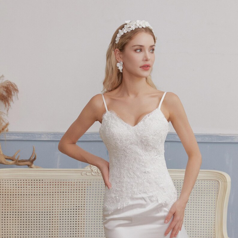 May include: A white wedding dress with a V-neckline and spaghetti straps. The bodice is adorned with intricate lace detailing. The model is wearing a floral headband and earrings. The dress has a satin skirt.
