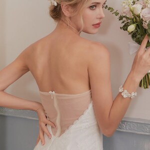 May include: A strapless white wedding dress with a sheer back panel and lace detailing. The bride is holding a bouquet of white and pink flowers and wearing a white floral bracelet. The dress features a delicate, romantic design.