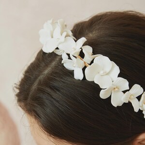 May include: A white floral headband featuring delicate, realistic-looking flowers with pearl accents. The flowers are arranged along a thin, gold-toned wire, creating an elegant and romantic hair accessory. Perfect for weddings or special occasions.