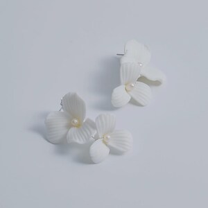 May include: A pair of white floral stud earrings. Each earring features a cluster of three or four petals with a small, round, pearl-like center. The earrings are set against a plain white background.