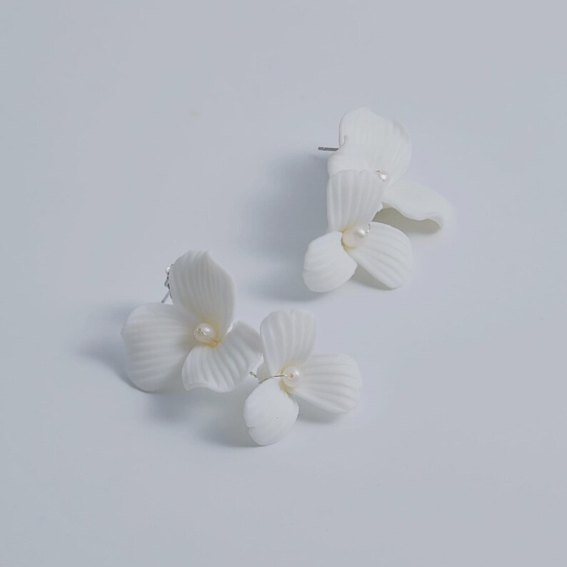 May include: A pair of white floral stud earrings. Each earring features a cluster of three or four petals with a small, round, pearl-like center. The earrings are set against a plain white background.