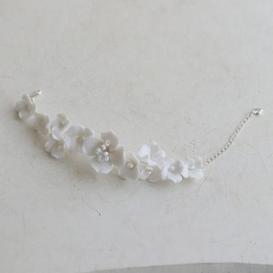 May include: A delicate white floral bracelet featuring multiple small, detailed flower blossoms. The bracelet is connected by a thin, silver chain. The flowers are clustered together, creating a graceful, elegant accessory.
