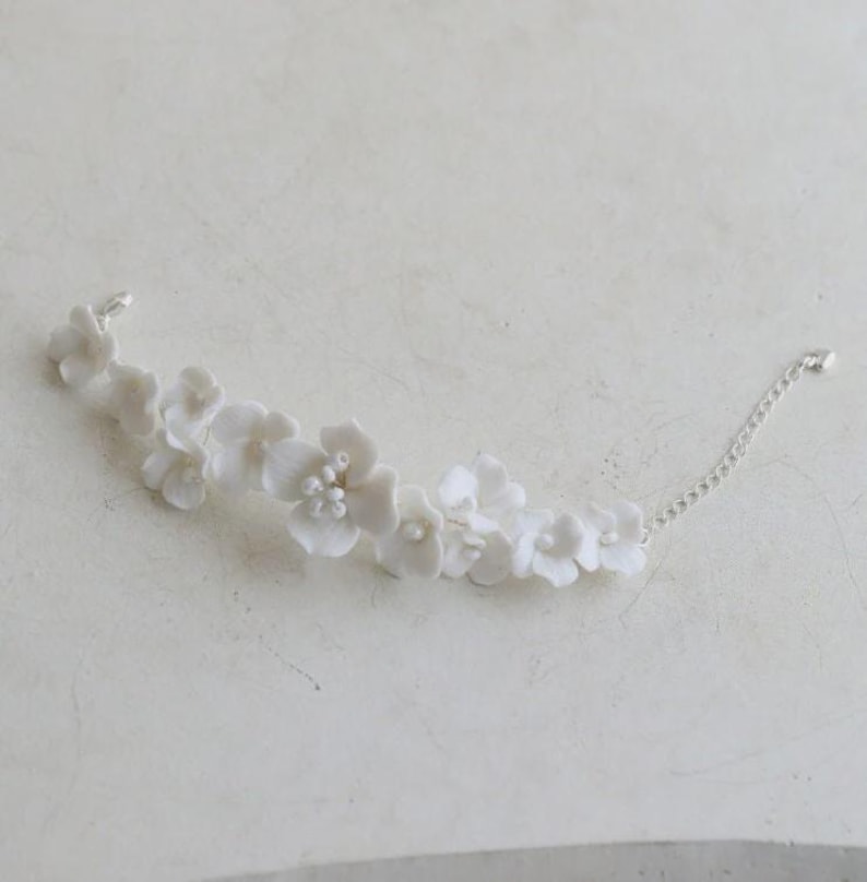 May include: A delicate white floral bracelet featuring multiple small, detailed flower blossoms. The bracelet is connected by a thin, silver chain. The flowers are clustered together, creating a graceful, elegant accessory.