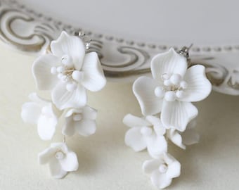 May include: A pair of white floral earrings. Each earring features multiple cascading white porcelain flowers with delicate detailing. The earrings are set against a soft, neutral background, highlighting their elegant design.