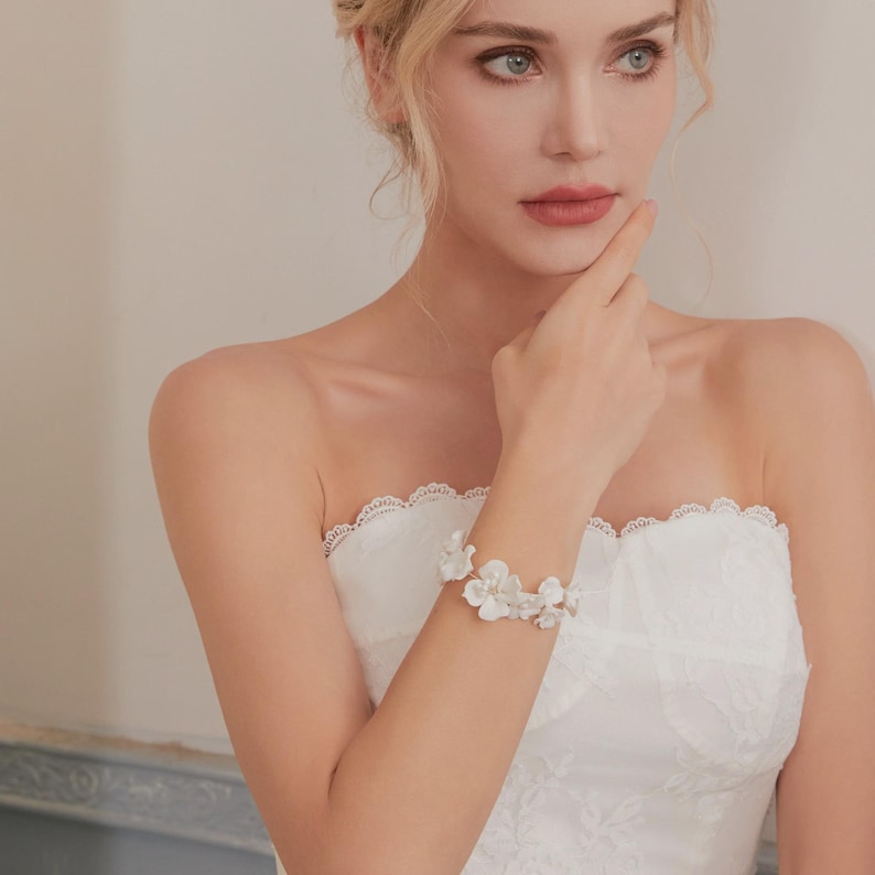 May include: A white floral bracelet adorns the wrist of a person wearing a strapless white dress. The bracelet features multiple white flower accents. The dress has a lace trim.