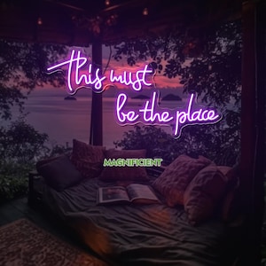 May include: A purple neon sign displaying the phrase "This must be the place" in a cursive font. The sign is set against a sunset over the sea. The word "MAGNIFICENT" is below the sign, with a bed and pillows in the foreground.