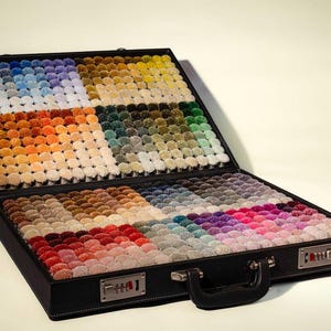 以下が含まれることがあります： さまざまな色のカーペットサンプルが入った、開いた黒いブリーフケース。サンプルはグリッドパターンに配置され、青と緑から赤と紫まで、幅広い色合いを示しています。ブリーフケースには黒いハンドルとコンビネーションロックが付いています。
