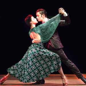 May include: A couple performing a tango dance. The woman wears a green patterned skirt and a matching shawl. The man is in a dark suit. They are in a close embrace, with the woman's red heels and the man's black shoes visible.