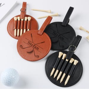 Könnte beinhalten: Braune und schwarze Golf-Tee-Halter aus Leder, jeweils mit Riemen und Schnalle. Die Halter sind rund und zeigen Golfschläger- und Ball-Designs mit personalisiertem Text. Golf-Tees und ein Golfball sind ebenfalls vorhanden.