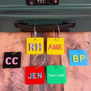 Puede incluir: Una colección de etiquetas de equipaje coloridas con varias iniciales y texto. Las etiquetas son cuadradas y están unidas a una maleta verde. Las etiquetas son amarillas, negras, rojas, azules y verdes, con lazos de metal plateado.