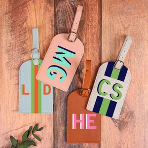 Op de afbeelding: Vier gepersonaliseerde bagagelabels in verschillende kleuren, waaronder lichtblauw, roze en bruin. Elke tag heeft initialen in vetgedrukte, contrasterende kleuren en een bijpassende riem. Geplaatst op een houten oppervlak.