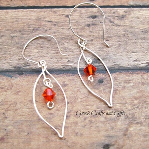 May include: Silver wire earrings with a teardrop shape and a small orange crystal bead hanging from each earring. The earrings are on a wooden surface.
