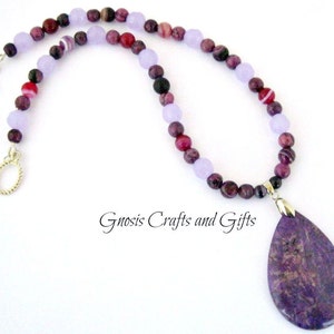 May include: A purple and lavender gemstone necklace with a large purple pendant. The necklace is made with various shades of purple and lavender beads, including some with white stripes. The pendant is a teardrop shape and has a marbled pattern.