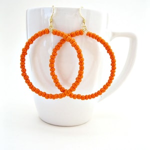 Peut inclure: Une paire de grandes boucles d'oreilles créoles orange faites de petites perles. Les boucles d'oreilles sont suspendues à une tasse blanche.