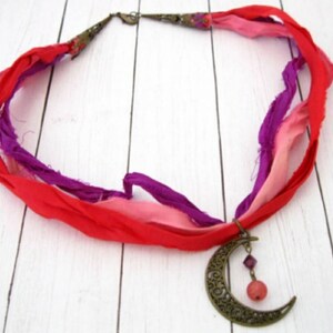 May include: A handmade necklace featuring a crescent moon pendant and colorful silk ribbons. The pendant is bronze-toned with intricate cut-out designs. The ribbons are red, pink, and purple, creating a bohemian style.