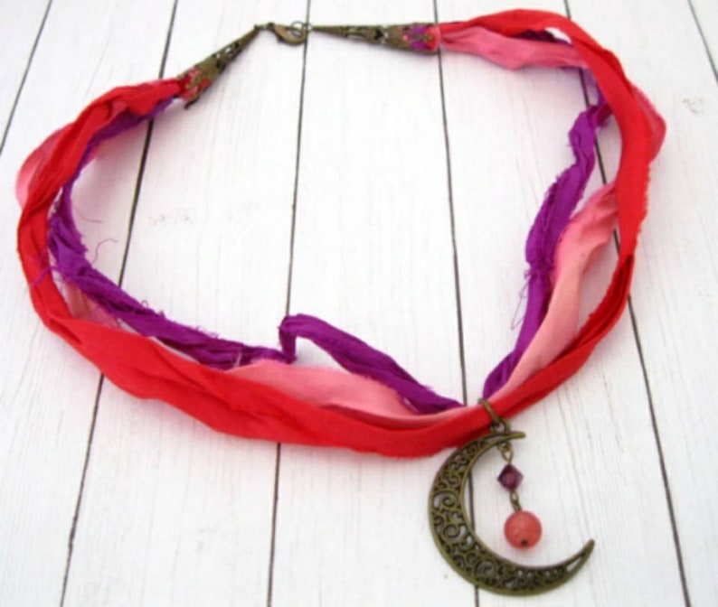 May include: A handmade necklace featuring a crescent moon pendant and colorful silk ribbons. The pendant is bronze-toned with intricate cut-out designs. The ribbons are red, pink, and purple, creating a bohemian style.