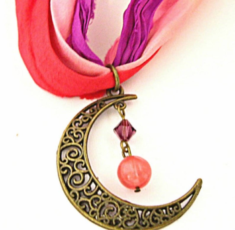 May include: A bronze-colored crescent moon pendant with a filigree design, suspended from a colorful ribbon. The pendant features a dangling pink crystal and a peach-colored bead. The ribbon is a mix of red, pink, and purple.