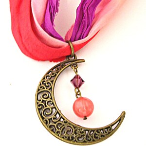 May include: A bronze-colored crescent moon pendant with intricate scrollwork, suspended from a colorful ribbon. The pendant features a dangling pink crystal and a round, peach-colored bead. The ribbon is a mix of pink, red, and purple.