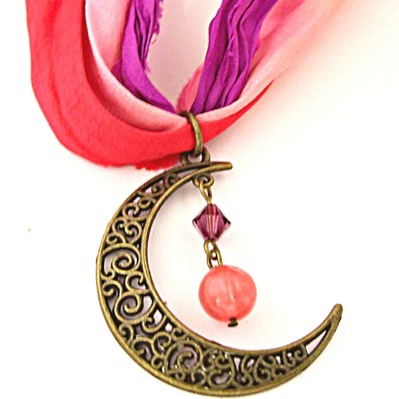 May include: A bronze-colored crescent moon pendant with intricate scrollwork, suspended from a colorful ribbon. The pendant features a dangling pink crystal and a round, peach-colored bead. The ribbon is a mix of pink, red, and purple.