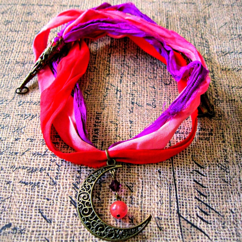 May include: A handmade necklace featuring a crescent moon pendant with a red bead and purple accents. The pendant hangs from a vibrant, multi-colored silk ribbon in shades of red, pink, and purple. The necklace is designed with an antique bronze finish.