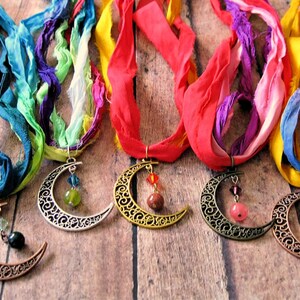 May include: A collection of handmade necklaces featuring crescent moon pendants with intricate designs. Each necklace has a colorful silk ribbon in shades of blue, green, purple, red, and yellow. The pendants are adorned with beads.