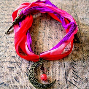 May include: A handmade necklace featuring a crescent moon pendant with intricate detailing, suspended from a vibrant red and purple silk ribbon. The pendant has a small red bead and other small beads hanging from it. The ribbon is tied in a loop.