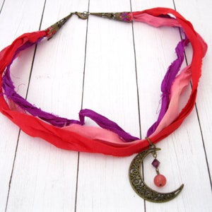 May include: A handmade necklace featuring a crescent moon pendant and colorful silk ribbons. The necklace has a bronze-colored moon pendant with a dangling pink bead and purple crystal. The ribbons are red, pink, and purple.