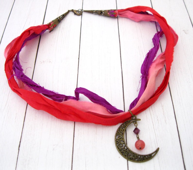 May include: A handmade necklace featuring a crescent moon pendant and colorful silk ribbons. The necklace has a bronze-colored moon pendant with a dangling pink bead and purple crystal. The ribbons are red, pink, and purple.