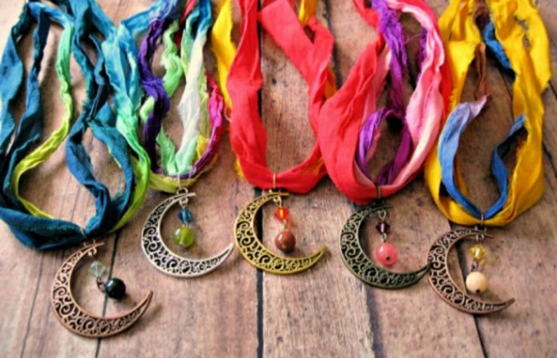 May include: Five handmade necklaces with crescent moon pendants and colorful silk ribbon. The pendants are in various metallic finishes and adorned with beads. The ribbons are in shades of blue, green, red, purple, and yellow.