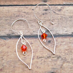 May include: Silver wire earrings with a teardrop shape and a small orange crystal bead hanging from each earring. The earrings are on a wooden surface.