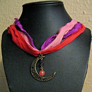 May include: A bronze-colored crescent moon pendant necklace with a red and pink gemstone and a multi-colored ribbon. The ribbon is red, pink, and purple. The necklace is displayed on a black mannequin.