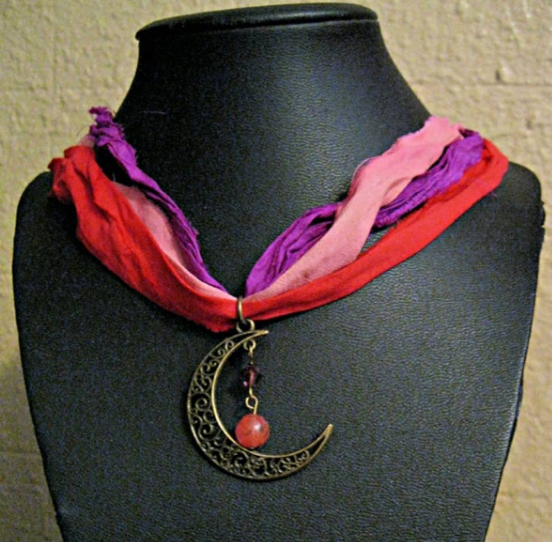 May include: A bronze-colored crescent moon pendant necklace with a red and pink gemstone and a multi-colored ribbon. The ribbon is red, pink, and purple. The necklace is displayed on a black mannequin.