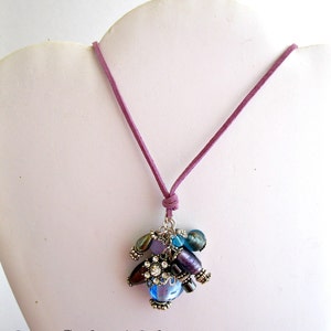 May include: A purple suede cord necklace with a charm featuring a cluster of blue, purple, and silver beads and a rhinestone accent.