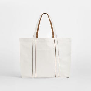 Puede incluir: Un bolso tote de lona blanca con asas y ribetes de cuero marrón. El bolso tiene forma rectangular con un bolsillo en la parte delantera. Las asas son lo suficientemente largas para llevarlo al hombro. Un bolso sencillo y práctico para el día a día.