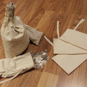 May include: Four white fabric bags, two are tied shut with a string, one is open and spilling out silver coins, and the other three are folded and stacked on top of each other.