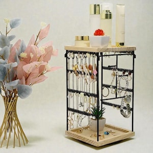 May include: A black metal jewelry organizer with a light wood base and top. The organizer displays necklaces, earrings, and bracelets. The top shelf holds skincare products and a small plant. A decorative floral arrangement is in the background.