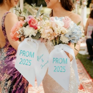 Könnte beinhalten: Zwei lächelnde Frauen halten Blumensträuße mit weißen Bändern. Auf den Bändern steht "PROM 2025" in türkisfarbenen Buchstaben, wobei ein Band auch "Alice & James" anzeigt. Die Sträuße zeigen rosa, weiße und blaue Blumen. Eine Frau trägt ein lila Blumenkleid.