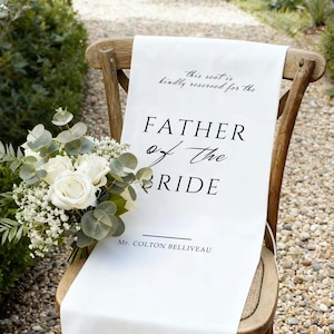 Puede incluir: Un camino de silla de tela blanca con el texto negro "FATHER of The BRIDE" está colocado sobre una silla de madera. Un ramo de rosas blancas y vegetación descansa sobre la silla. El texto "Mr. Colton Belliveau" también está impreso.