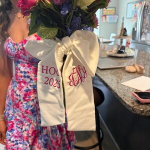 May include: A bouquet of flowers with a white ribbon. The ribbon has the text "HO 2025" in red, along with a monogram. The flowers include pink, purple, and white blooms.