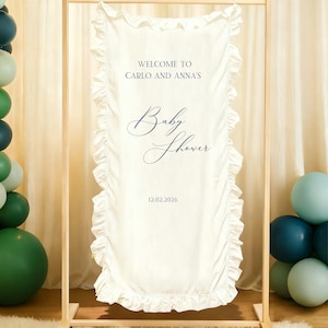 May include: Cream-colored baby shower banner with ruffled edges and vertical stripes. The banner reads "WELCOME TO CARLO AND ANNA'S" and "Baby Shower" in elegant navy blue script, with the date 12.02.2026 below. The banner is displayed on a wooden frame.