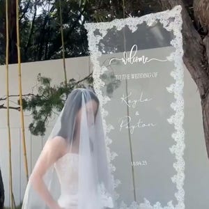 Pode incluir: Uma placa de casamento de acr&iacute;lico transparente com uma borda de renda branca e uma escrita elegante que diz "Bem-vindos ao casamento de Kayla & Payton 10.04.25". A placa &eacute; exibida ao ar livre perto de uma &aacute;rvore.