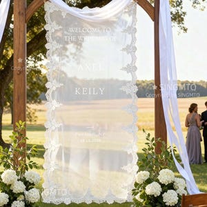 May include: A wooden wedding arch with a sheer white fabric panel featuring the text "WELCOME TO THE WEDDING OF AXEL and KEILY" and the date 18.10.2025. The arch is decorated with white draping fabric and floral arrangements.